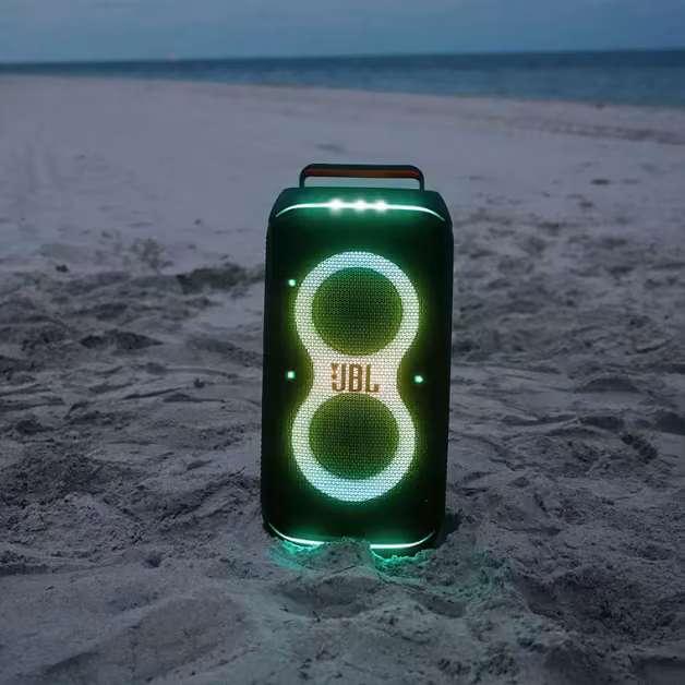 A JBL portable speaker with glowing green and yellow lights sits on a sandy beach at dusk, ocean and clouds in the background.