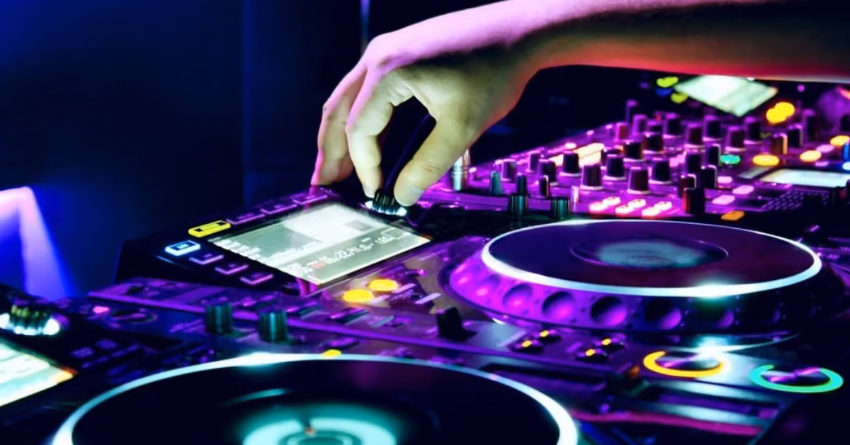 A DJ’s hand adjusts a knob on a brightly lit mixer and turntable at a lively party, with colorful lights in the background.