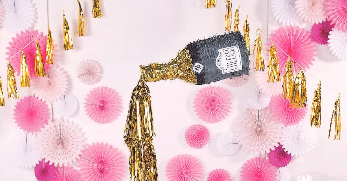 Champagne bottle piñata pours gold streamers, with pink and white fans and gold tassels for a festive party backdrop.