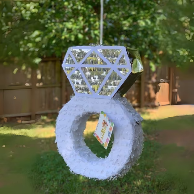 A diamond ring piñata with a shiny silver top and white band hangs outdoors, with green foliage and a wooden fence behind.