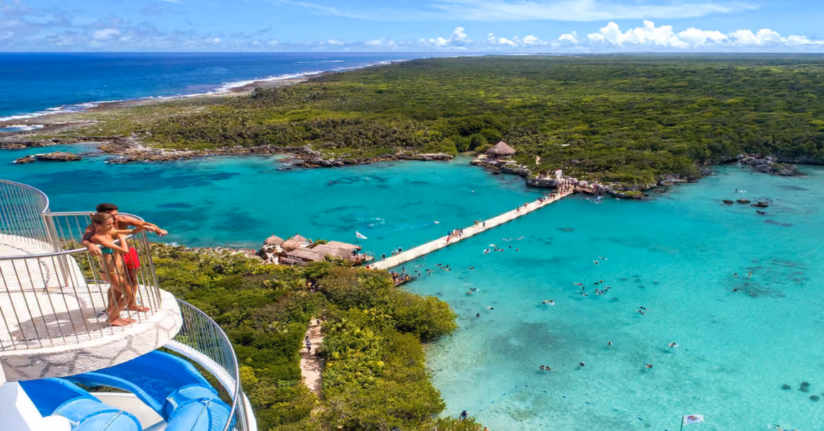 A couple stands on a water slide platform at Xel-Ha, overlooking a turquoise lagoon, lush greenery, and swimmers below.