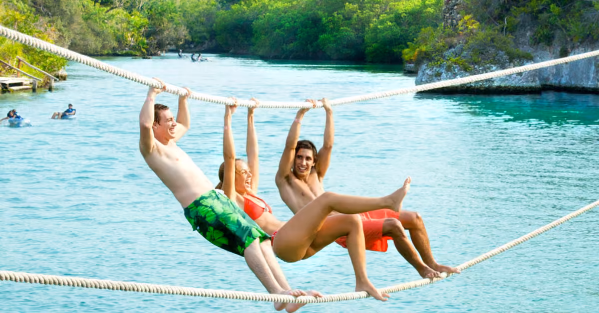 Three friends in swimsuits laugh, swinging from ropes over a turquoise river, greenery and swimmers in the background.
