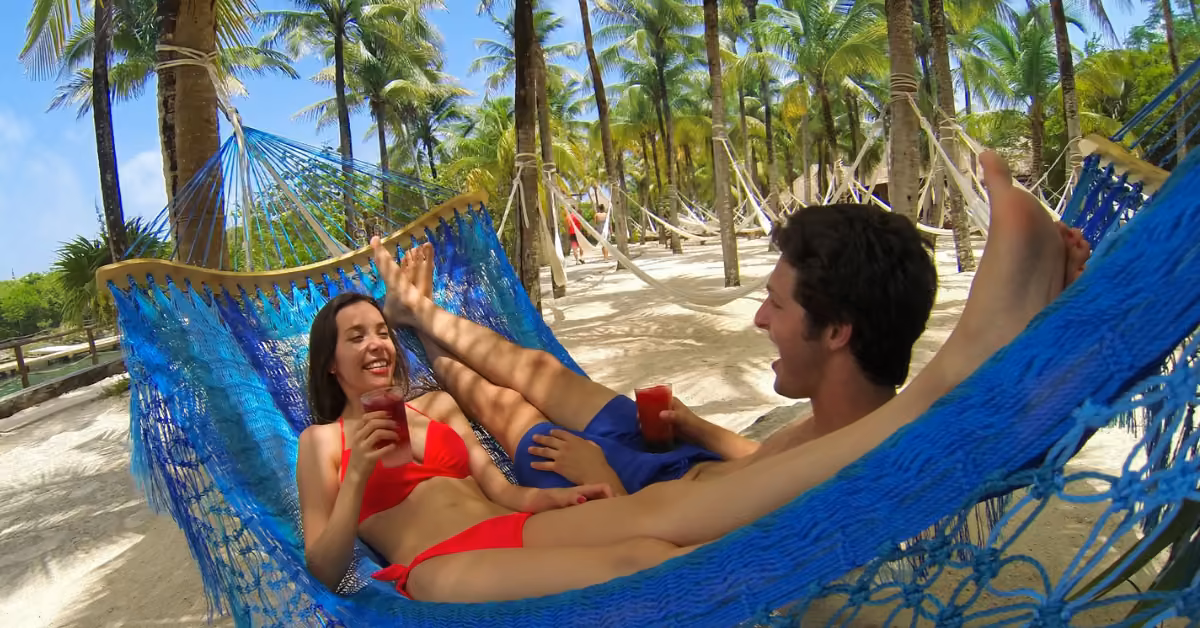 Two people smile and hold drinks while relaxing in a blue hammock on a sunny tropical beach, surrounded by palm trees.
