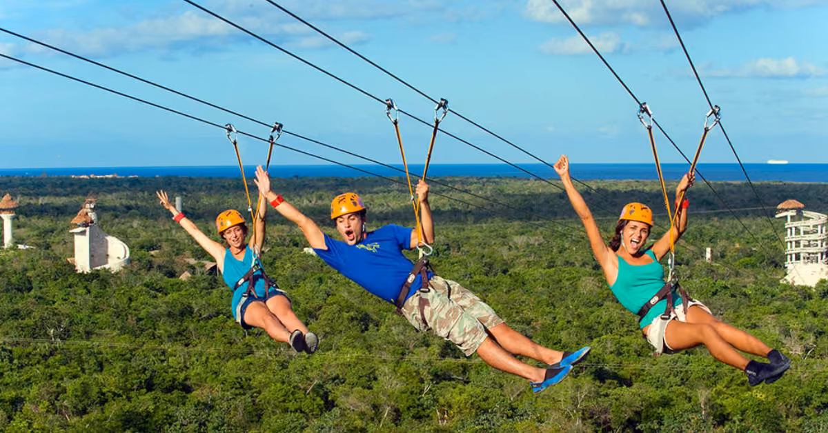 Three people in helmets and harnesses ride ziplines side by side over a green forest, smiling and raising their arms excitedly.