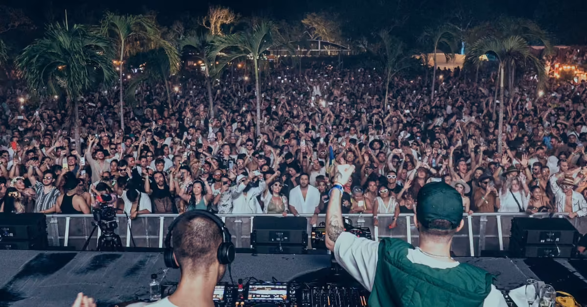 A lively crowd dances under palm trees and lights at Zamna Music Festival as two DJs perform on stage at night.