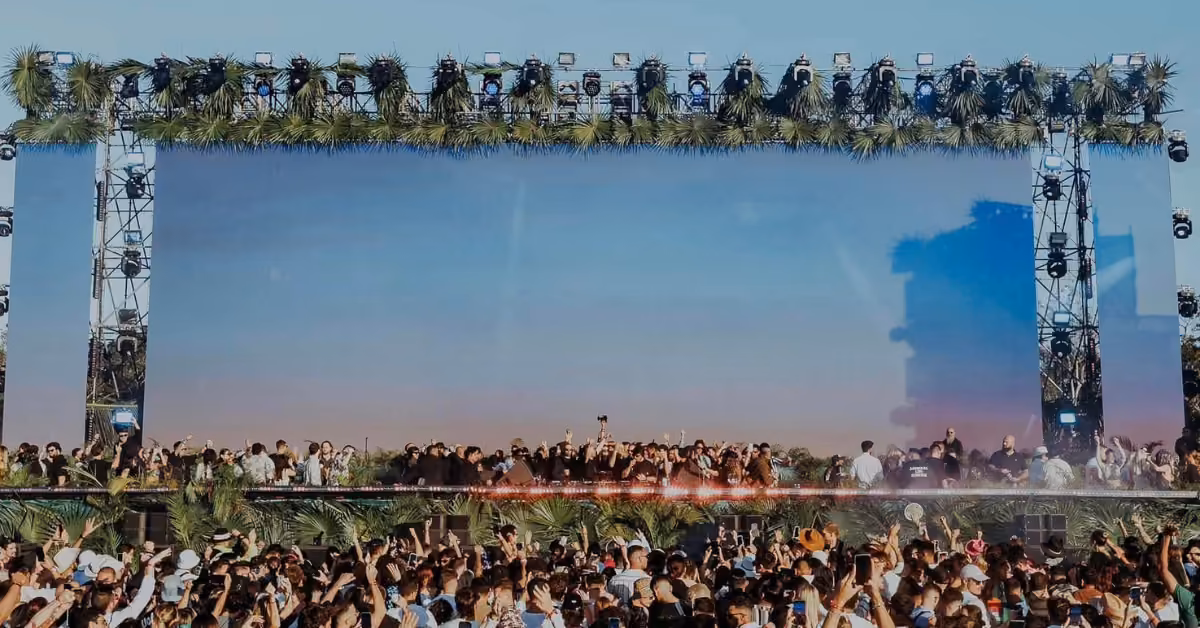 A crowd enjoys the Zamna Music Festival at sunset, raising hands in front of a wide stage with palm leaf decorations.