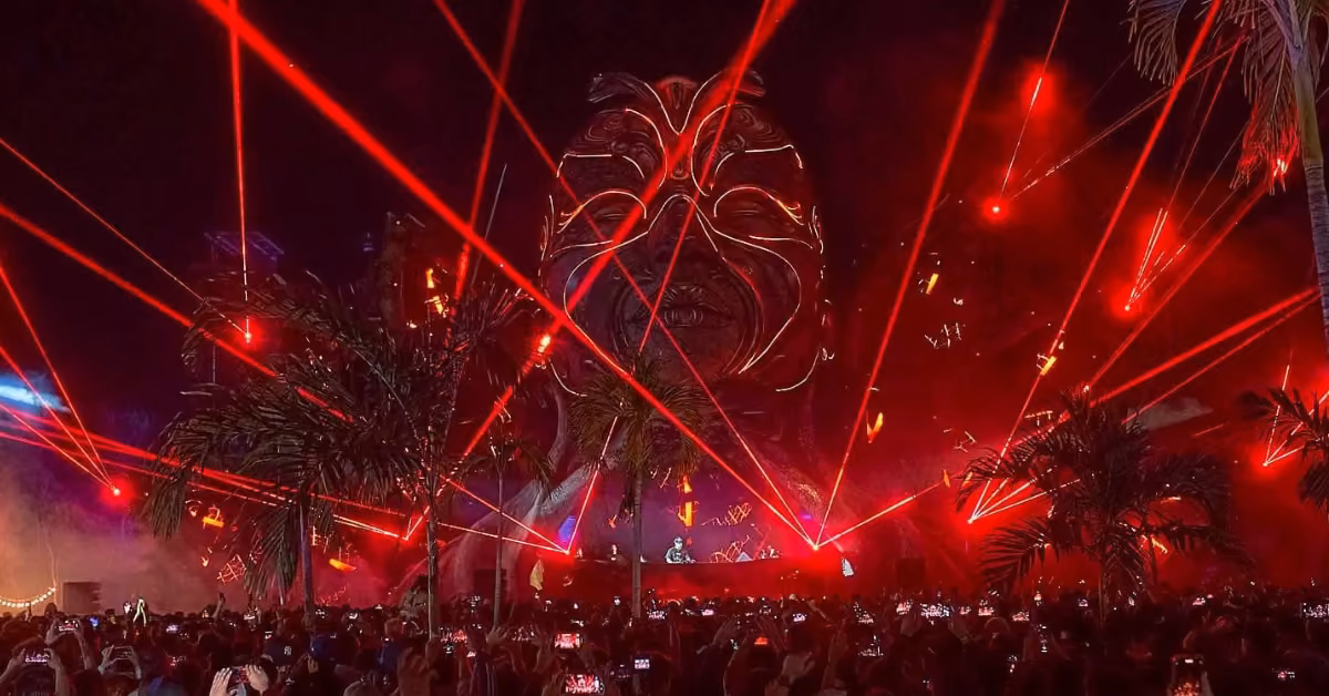 Nighttime Zamna Festival: large crowd, red lasers, palm trees, and a glowing tribal mask behind the DJ. Fans record on phones.