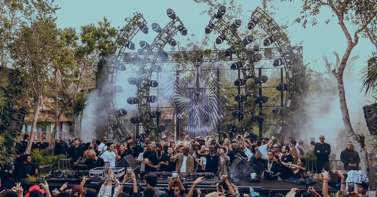 Crowds dance by a forested stage as a DJ plays, sunlight shining through trees at an energetic daytime music festival.