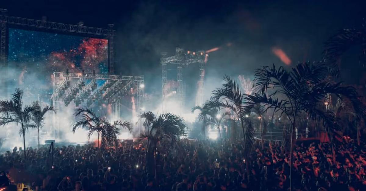 Concertgoers gather under palm trees at night, enjoying music amid bright stage lights and a vibrant, colorful screen.