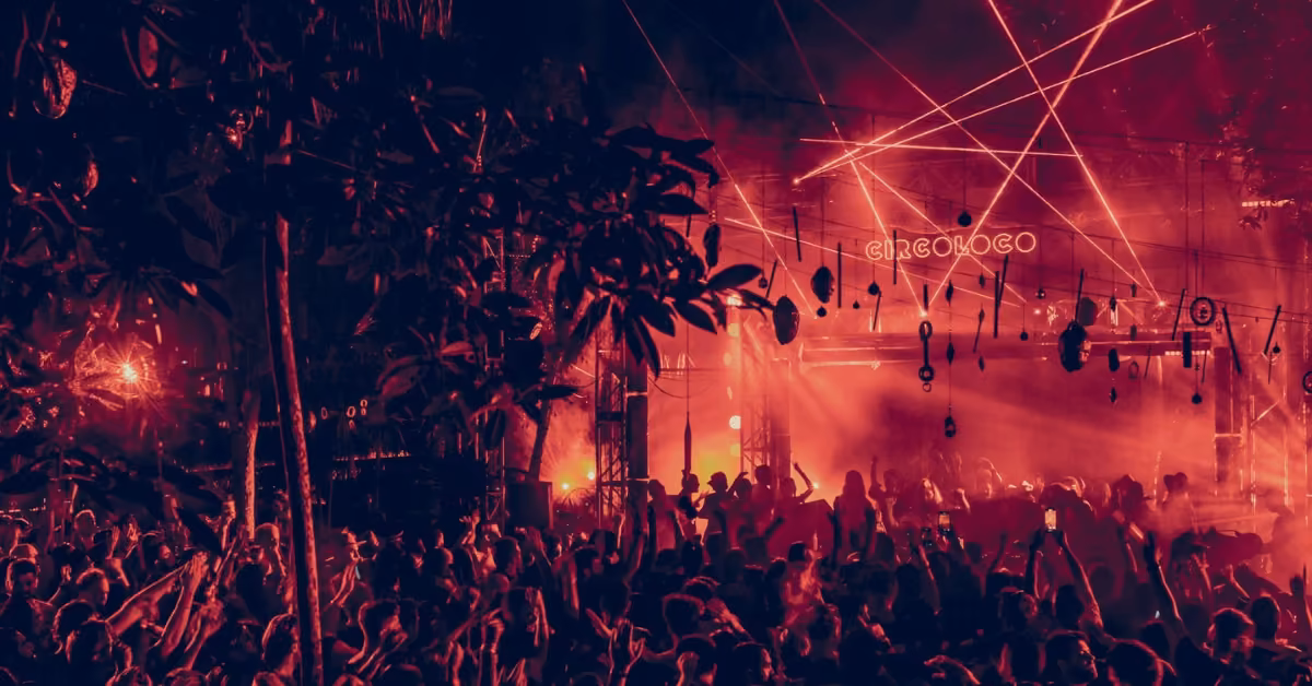 A lively crowd at an outdoor night music festival with red lights, lasers, fog, trees, and a stage sign reading CIRCOLOCO.