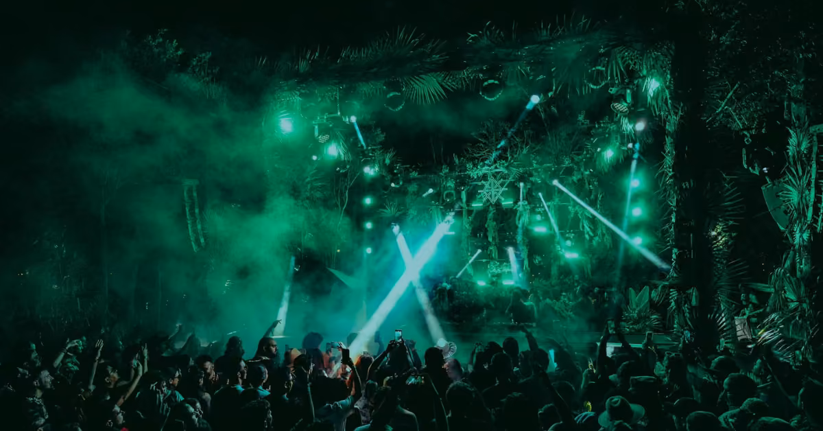 Crowds enjoy a nighttime music festival, green lights and fog highlighting palm-decorated stages at Zamna Music Festival.