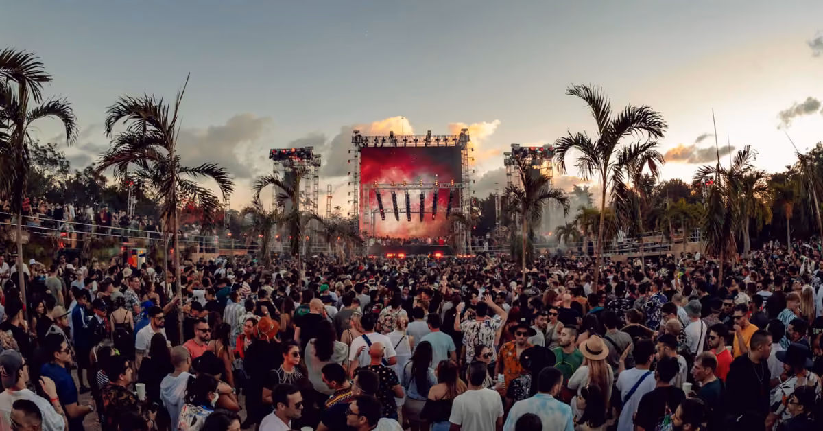 A crowd enjoys an outdoor music festival by palm trees, watching a brightly lit stage at sunset beneath a partly cloudy sky.