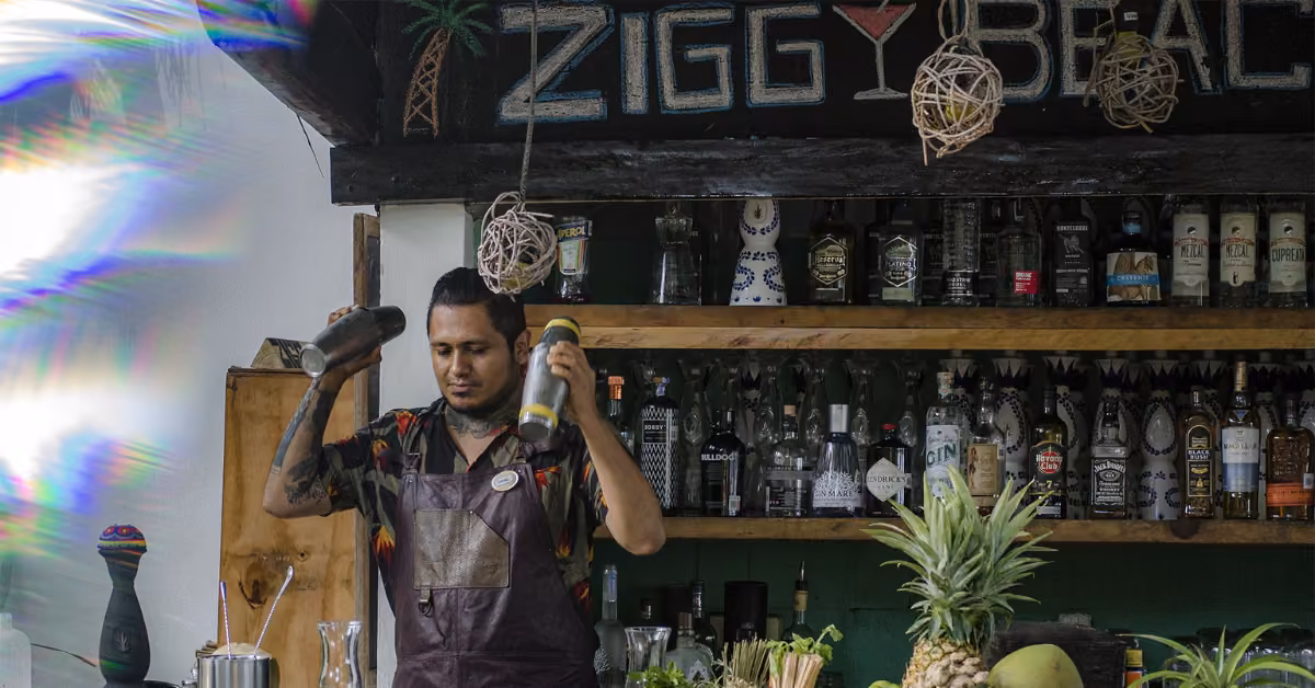 A bartender stands behind the bar at Ziggy’s Beach Club, shaking two cocktail shakers amid bottles and tropical fruits. A sign above reads “Ziggy Beach,” adding to the lively, tropical atmosphere.