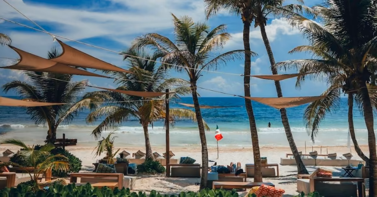 A tropical beach scene at Ziggy’s Beach Club with palm trees, lounge chairs, and triangular shade sails. The turquoise ocean has gentle waves, and a small Mexican flag is visible near the water under a partly cloudy sky.