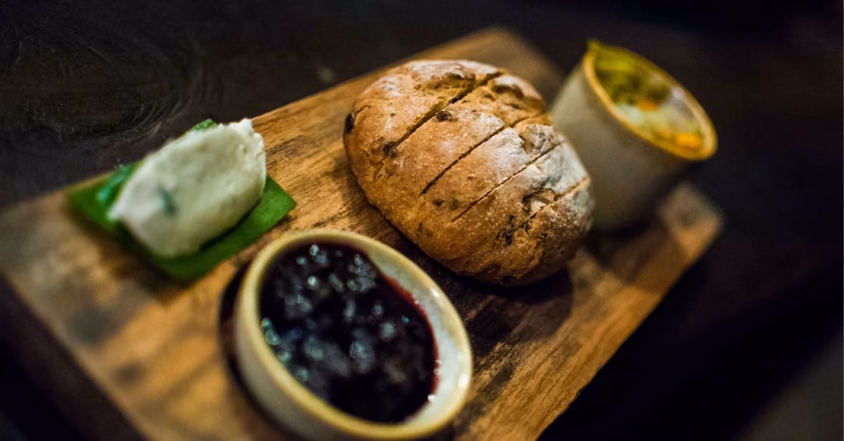 A wooden board at Ziggy’s Beach Club holds a round loaf of bread, a small dish of dark jam, soft white cheese on a green leaf, and a small bowl with a baked dish, all arranged neatly.