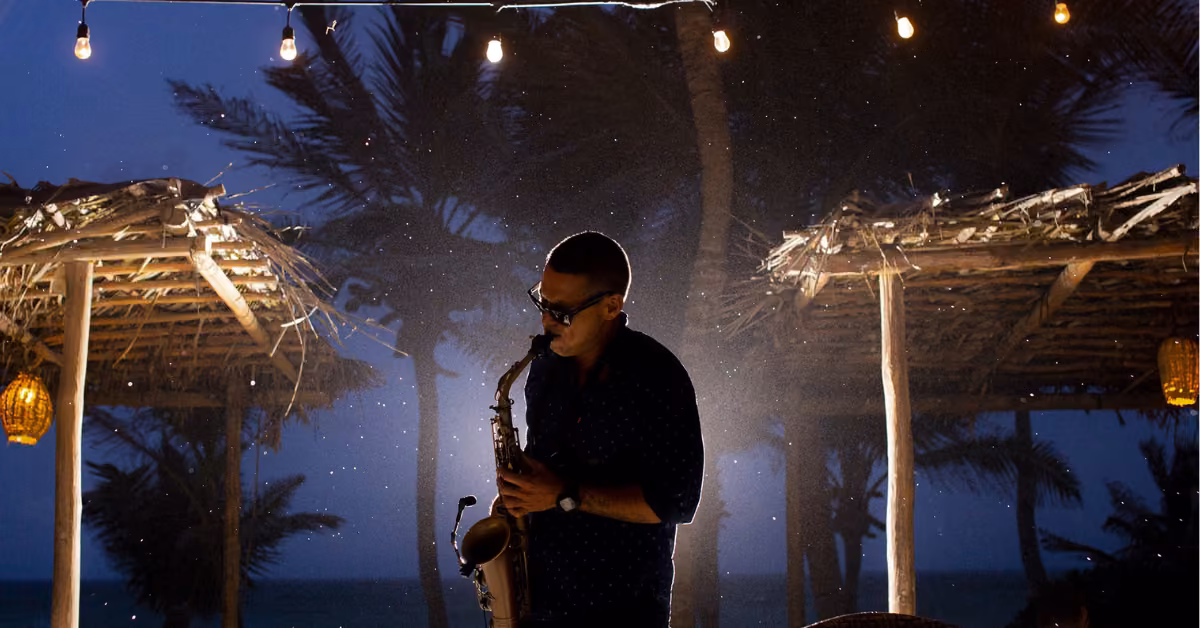 A man plays the saxophone under string lights at night at Ziggy’s Beach Club, surrounded by palm trees and rustic wooden shelters, with the ocean visible in the background.