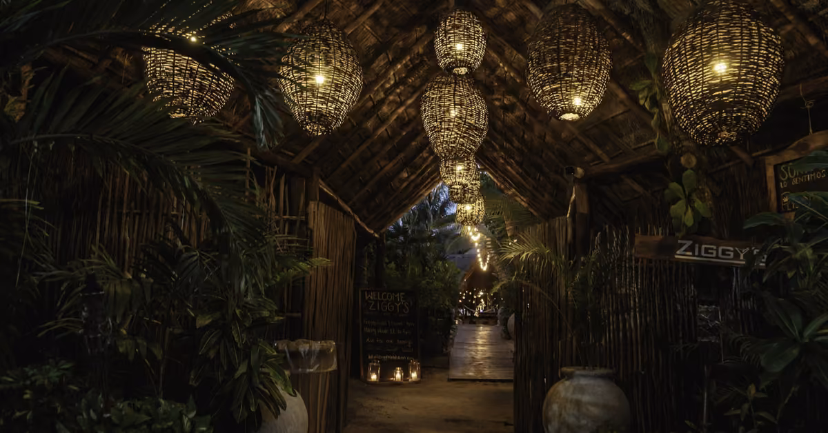 A dimly lit, tropical entrance at Ziggy’s Beach Club features woven lanterns hanging from a thatched roof, surrounded by lush plants and large ceramic pots, creating a warm, inviting ambiance leading to the restaurant.