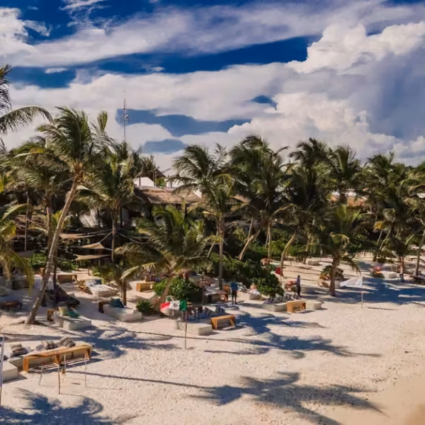 Luxurious beachside lounge beds and umbrellas await on white sand at one of the area’s exclusive beach clubs, surrounded by tall palm trees, with a resort and dramatic, cloud-filled blue sky in the background.