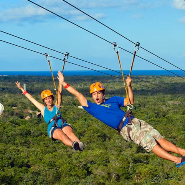 Two people ziplining side by side over a green forest with blue sky and ocean behind them, arms outstretched in excitement.