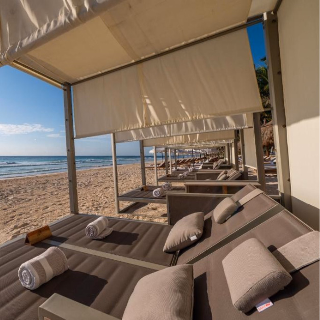 A row of shaded beach cabanas with cushioned lounge chairs and rolled towels overlooks a sandy beach and the ocean under a clear blue sky.