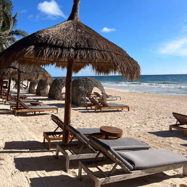 Wooden lounge chairs with cushions sit under large thatched umbrellas on a sandy beach. The ocean waves are visible in the background under a blue sky, and palm trees are partly visible to the left.