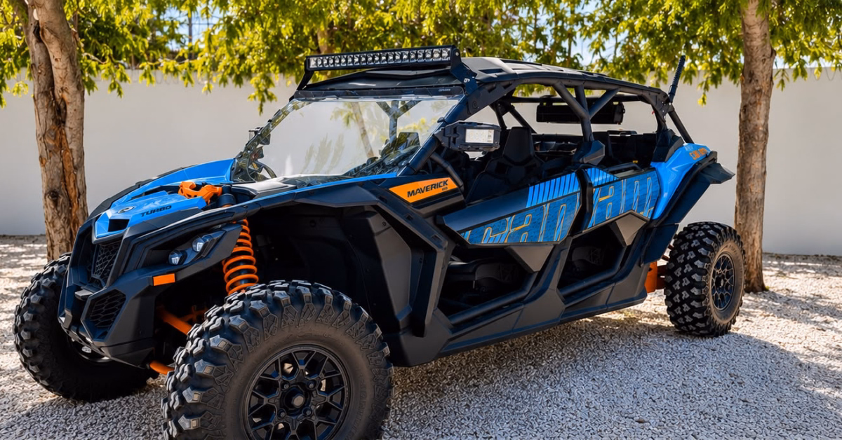 A blue and black ATV with large tires is parked on gravel under two trees, near a white wall and greenery in the background.