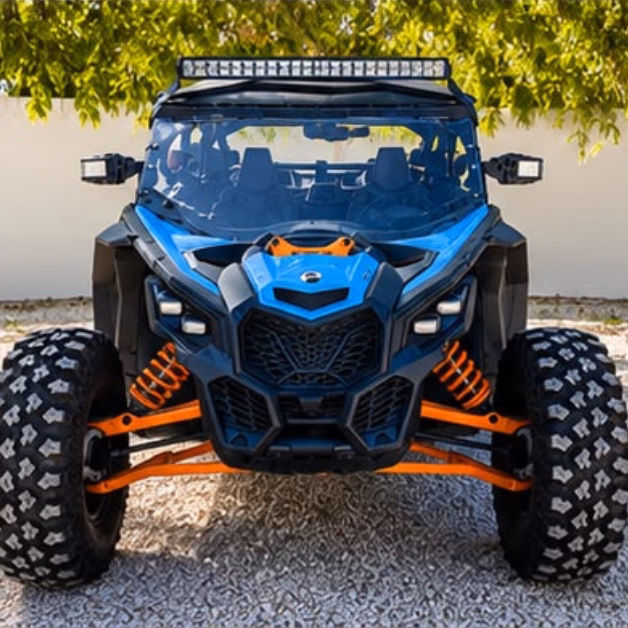 A blue and black ATV with large tires and orange suspension is parked on gravel, with green trees and a white wall behind it.