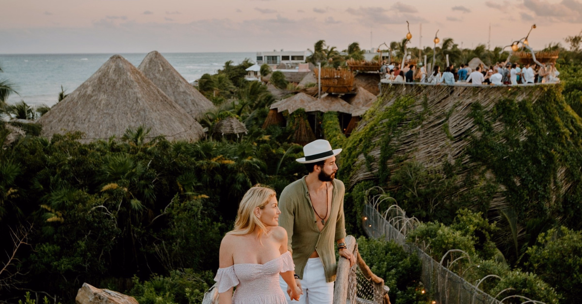 A couple dressed in light, summery clothing walks hand in hand along a pathway surrounded by lush greenery, with thatched-roof structures and a group gathered on the kin-toh rooftop near the ocean.