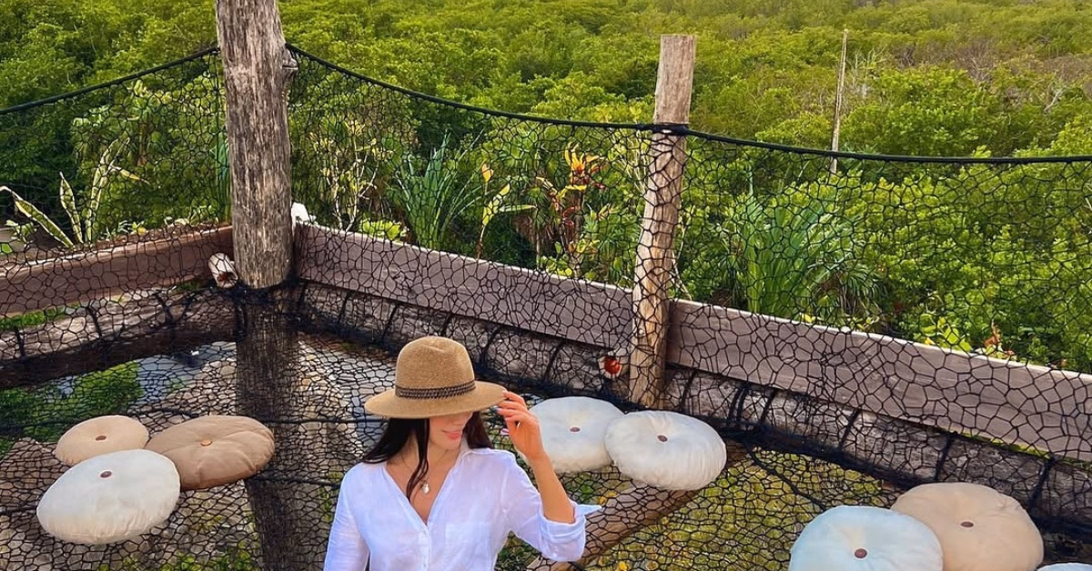 A woman in a white shirt and tan hat sits on a netted platform with round cushions at the kin-toh rooftop, surrounded by lush green trees and wooden posts in an inviting outdoor setting.
