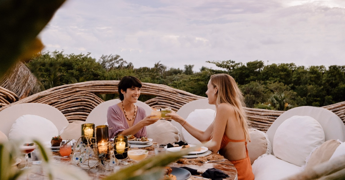 Two women sit at an outdoor table with food and drinks, smiling and clinking glasses at a kin-toh rooftop. Woven furniture, cushions, and candlelight create a cozy vibe amid lush greenery under a cloudy sky.