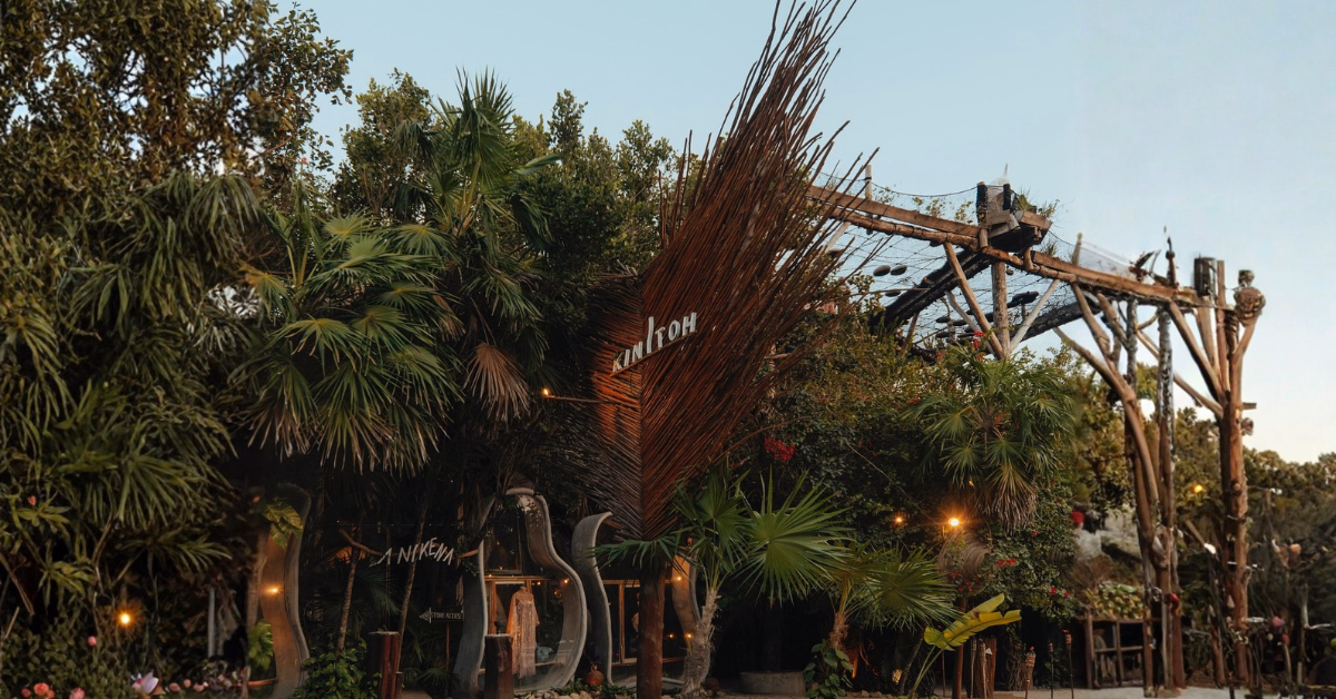 Outdoor entrance to a tropical-themed establishment surrounded by lush palm trees and greenery, featuring wooden art structures, hanging lights, and a wooden archway reminiscent of a kin-toh rooftop ambiance set against a clear sky.