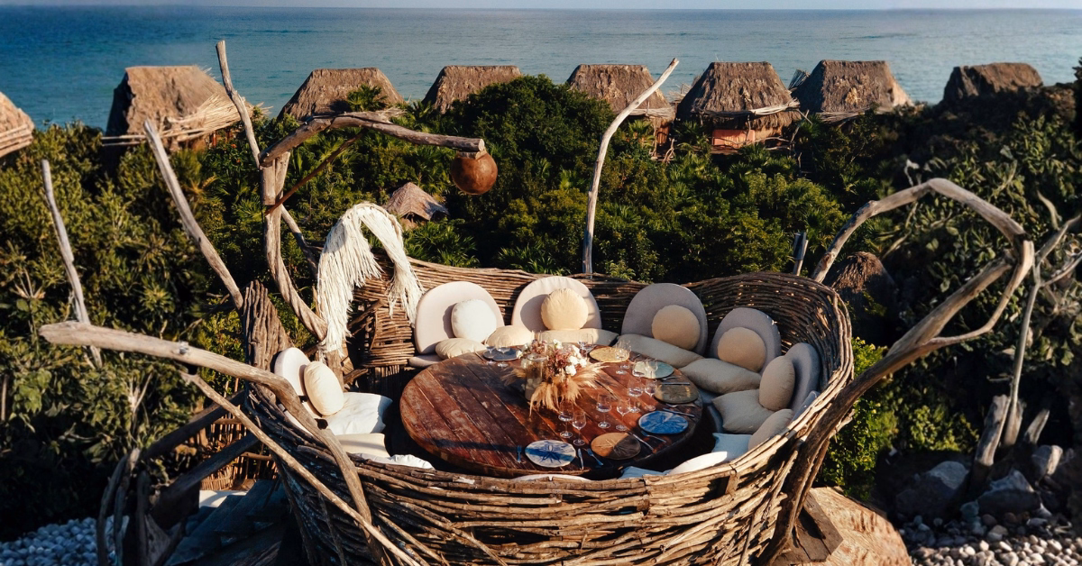 A large, nest-like wooden seating area with round cushions and a table sits above lush greenery on the kin-toh rooftop, with rustic thatched-roof huts and the ocean visible in the background.
