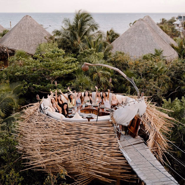 A group of people sit in a circular, nest-shaped outdoor lounge on the Kin-Toh rooftop, elevated among lush greenery with thatched-roof huts and the ocean in the background. Everyone appears to be celebrating or posing for a photo.