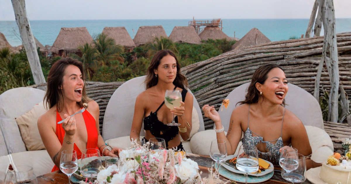 Three women dressed in elegant outfits sit at an outdoor table on the Kin-Toh rooftop, enjoying food and drinks surrounded by flowers and plates. Behind them are tropical huts, lush greenery, and a stunning ocean view.