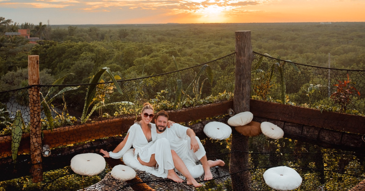 A couple wearing white robes sits on a netted platform with cushions at the Kin-Toh rooftop, surrounded by greenery and trees, enjoying a stunning sunset view over the forest.