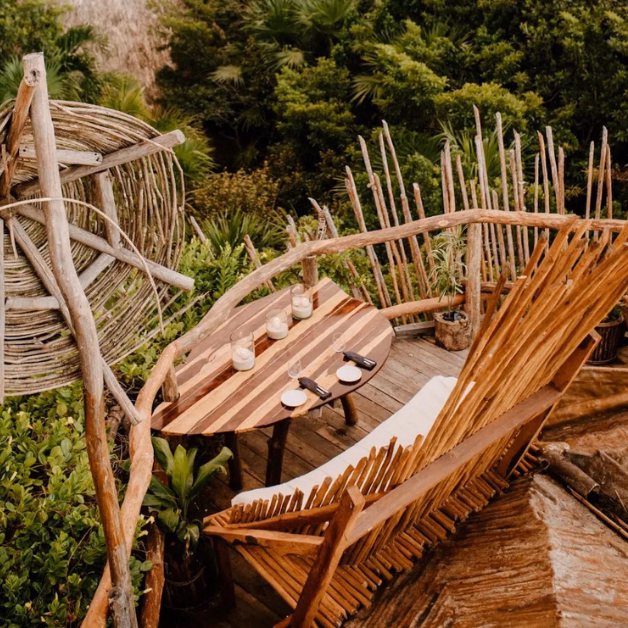A rustic outdoor seating area reminiscent of the kin-toh rooftop, featuring a wooden table set with four glasses and a small bowl, surrounded by wooden chairs and natural branches, with lush greenery in the background.
