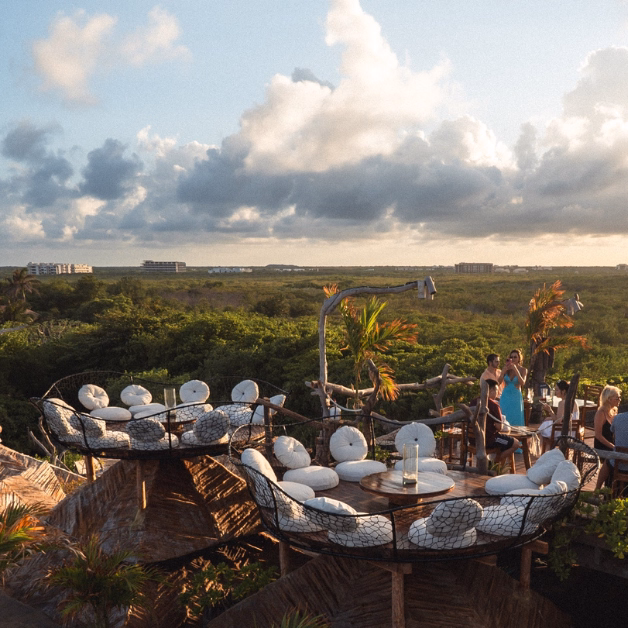Rooftop lounge with round tables and chairs overlooks greenery at sunset; people relax and enjoy the scenic view.
