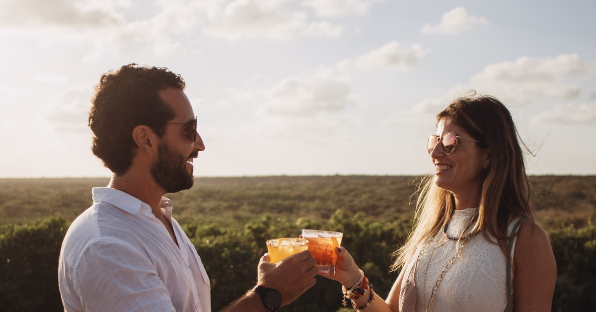 A man and a woman wearing sunglasses smile and clink drinks outdoors on a tseen-ja rooftop, with a scenic, sunny landscape and blue sky in the background.