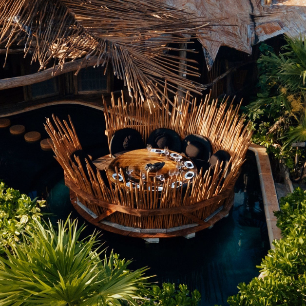 A round wooden dining table with black chairs sits in a nest-like structure made of sticks on the tseen-ja rooftop, surrounded by lush greenery and water, creating a tropical, nature-inspired dining setting.