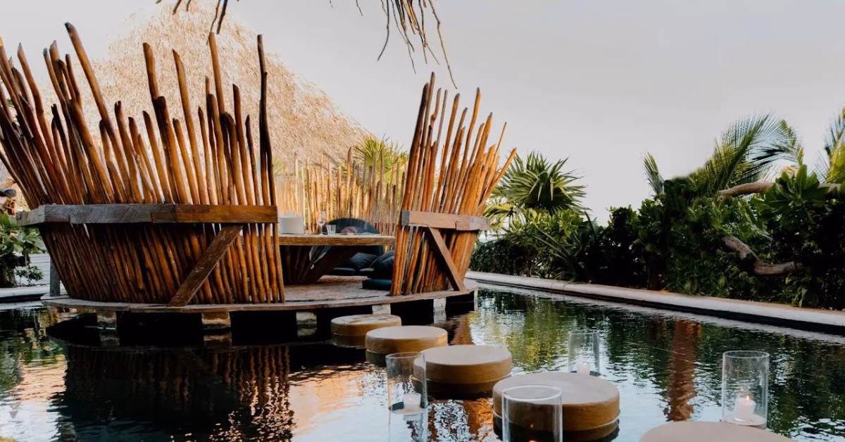 A round wooden seating area with tall vertical slats stands over water, accessible by circular stepping stones. Green plants and a thatched roof structure evoke the tranquil ambiance of a tseen-ja rooftop. Glass candle holders sit on the water's edge.