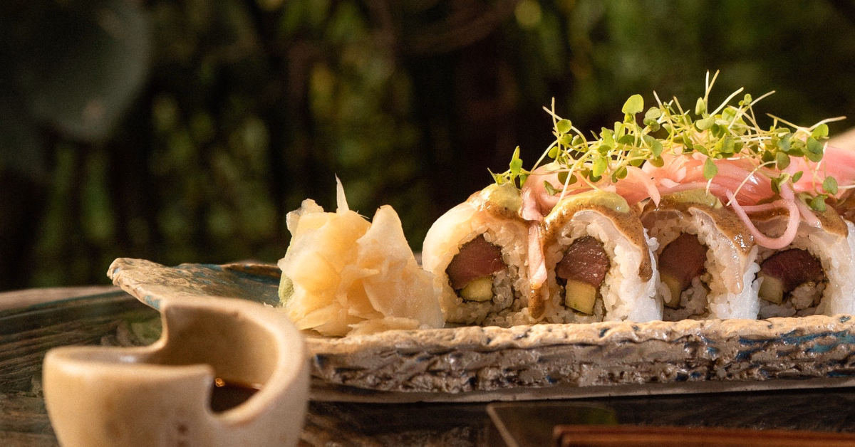 A close-up of sushi rolls topped with microgreens and pink pickled onions, served on a rustic ceramic plate at tseen-ja rooftop, with pickled ginger and soy sauce, set against a blurred green background.