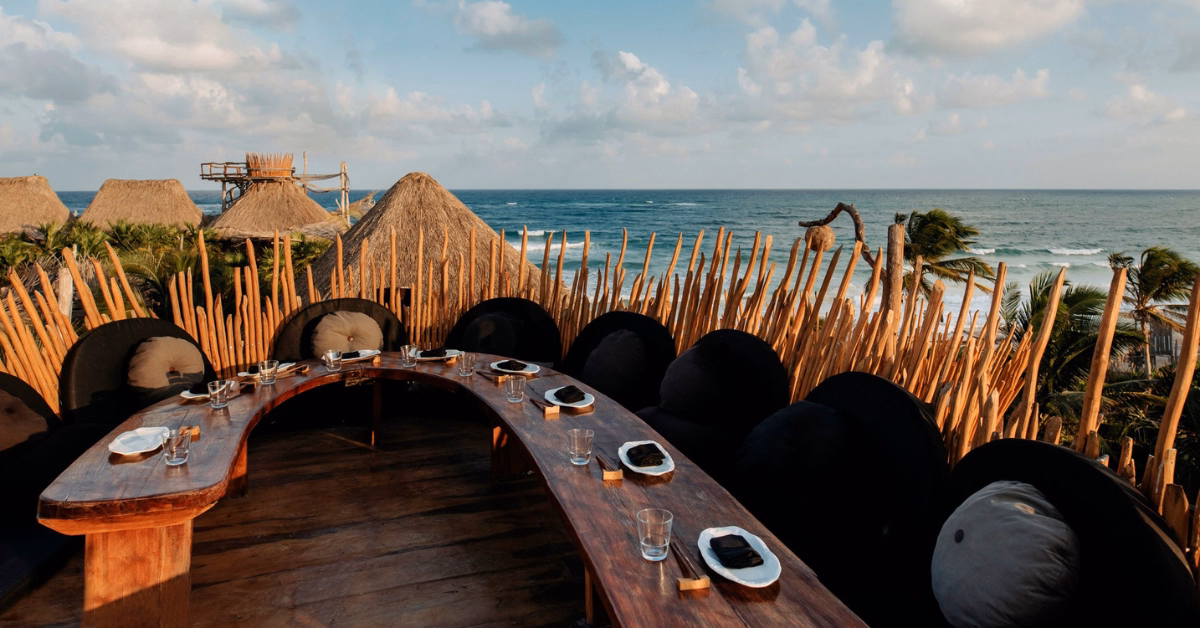 Curved wooden table with place settings on a tseen-ja rooftop terrace, surrounded by round black cushions and rustic fencing, overlooking palm trees, thatched roofs, and the ocean under a partly cloudy sky.
