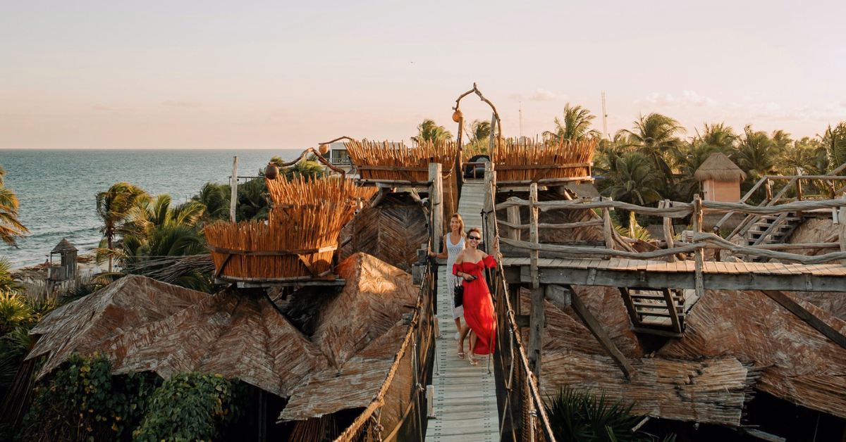 Two people walk on a wooden bridge among rustic, treehouse-style structures near the ocean and tseen-ja rooftop, surrounded by palm trees at sunset. One person wears a flowing red dress.