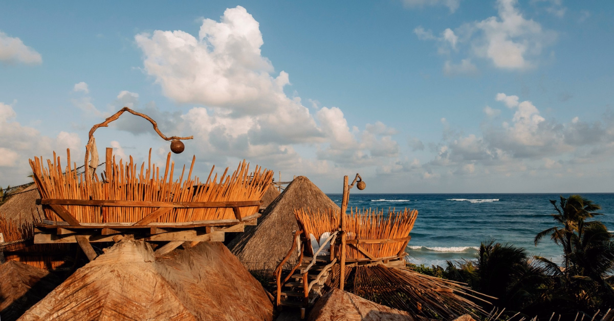 Thatched-roof structures with decorative wooden fences and traditional tseen-ja rooftop accents overlook a tropical beach and the ocean. Palm trees sway as waves roll in beneath a partly cloudy blue sky.