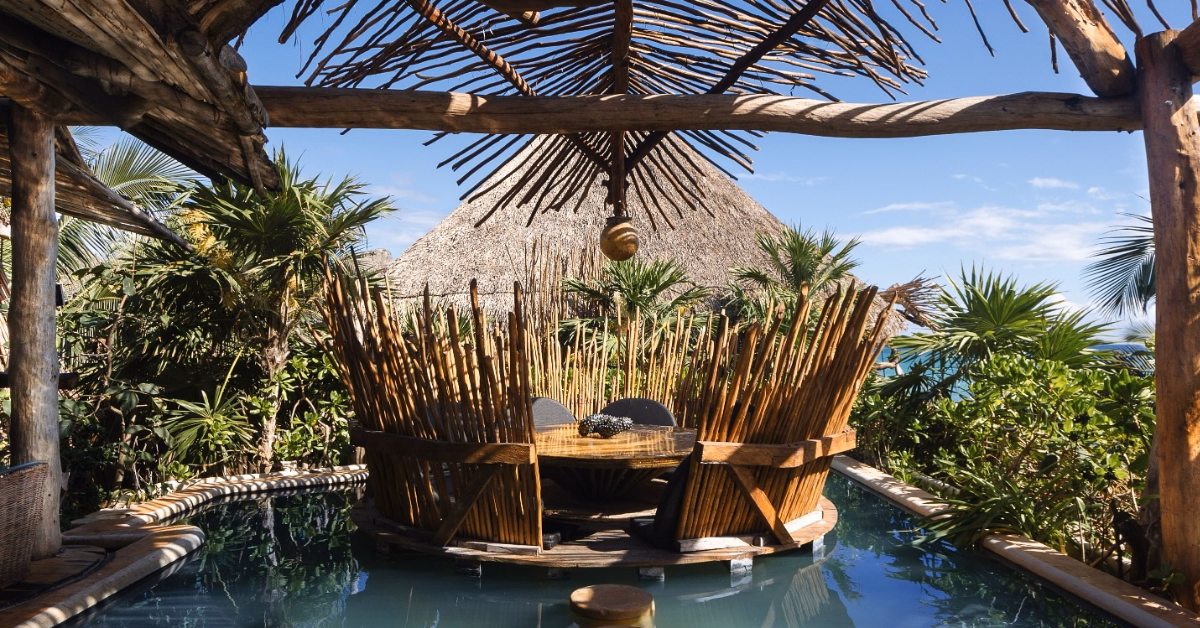 A rustic wooden dining table with high, spiky wooden chairs sits on a tseen-ja rooftop deck above a pool, surrounded by tropical plants and shaded by a thatched roof. Blue sky and ocean complete the serene backdrop.