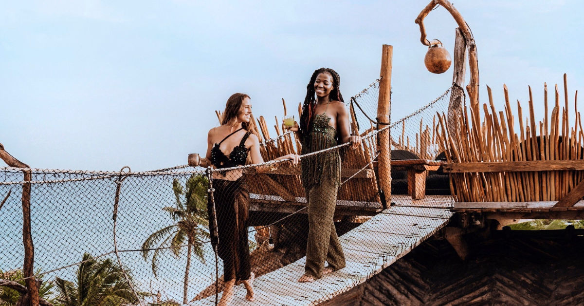 Two women in summer outfits walk across a wooden suspension bridge holding drinks, with palm trees, the ocean, and the vibrant energy of tseen-ja rooftop visible in the background on a sunny day.