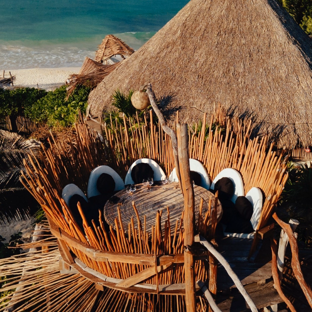 Aerial view of a rustic tseen-ja rooftop outdoor dining area with a round table and oval-backed chairs, surrounded by wooden sticks, beneath a large thatched roof by the ocean, with sand and greenery in the background.