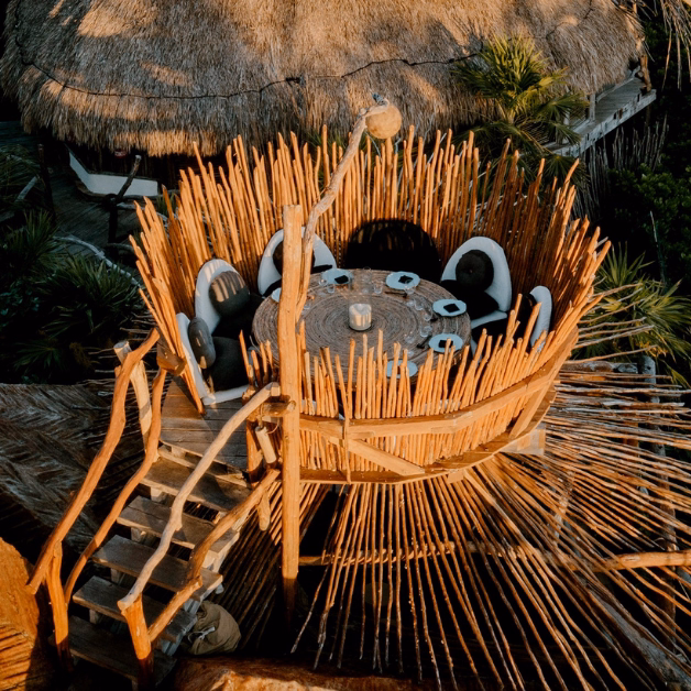A round, elevated dining table on the tseen-ja rooftop is surrounded by tall wooden sticks like a bird’s nest, with curved chairs and a wooden staircase, set among palm trees and thatched roofs.