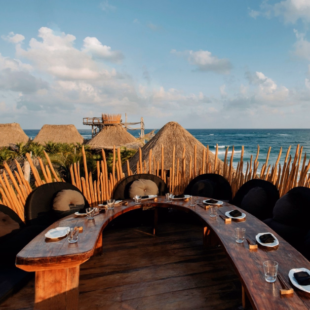 A curved wooden dining table set with plates and glasses overlooks the ocean from a tseen-ja rooftop, surrounded by thatched-roof huts and wooden fence posts under a blue sky with scattered clouds.