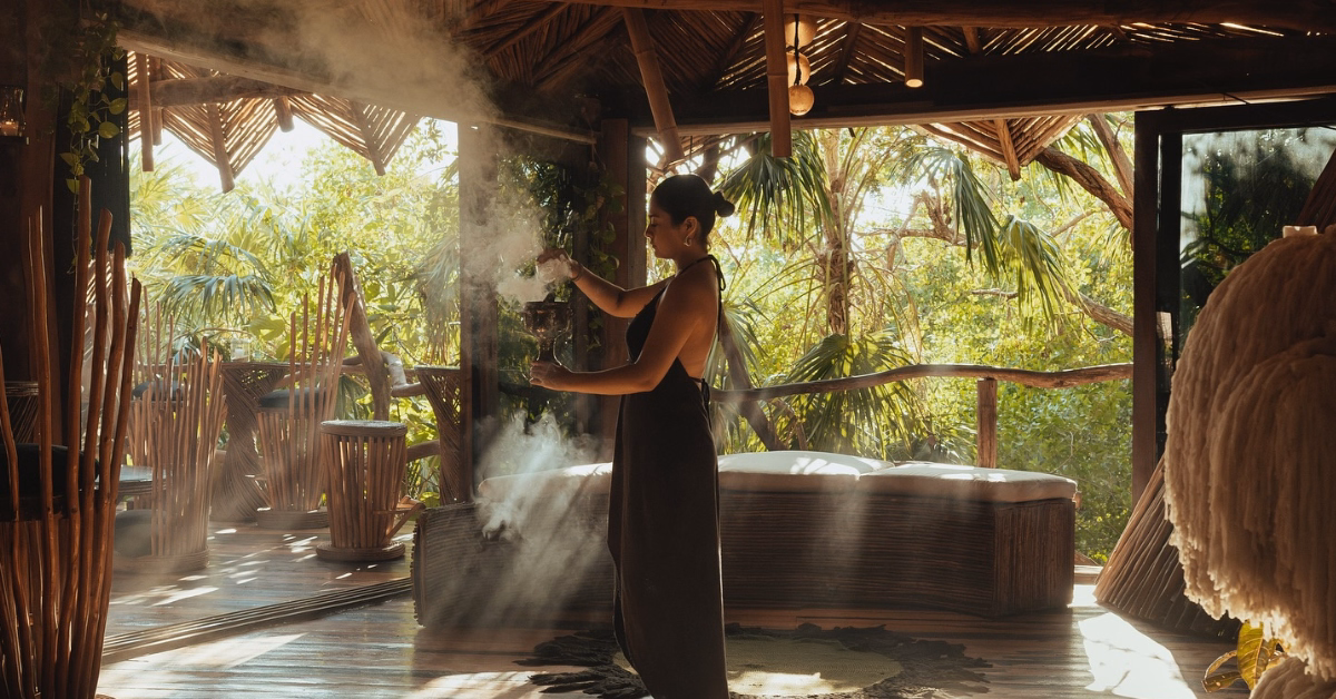 A woman in a dark dress stands in a sunlit, rustic wooden room on a tseen-ja rooftop, surrounded by greenery and holding a smoking bundle as sunlight filters through, creating a serene and tranquil atmosphere.